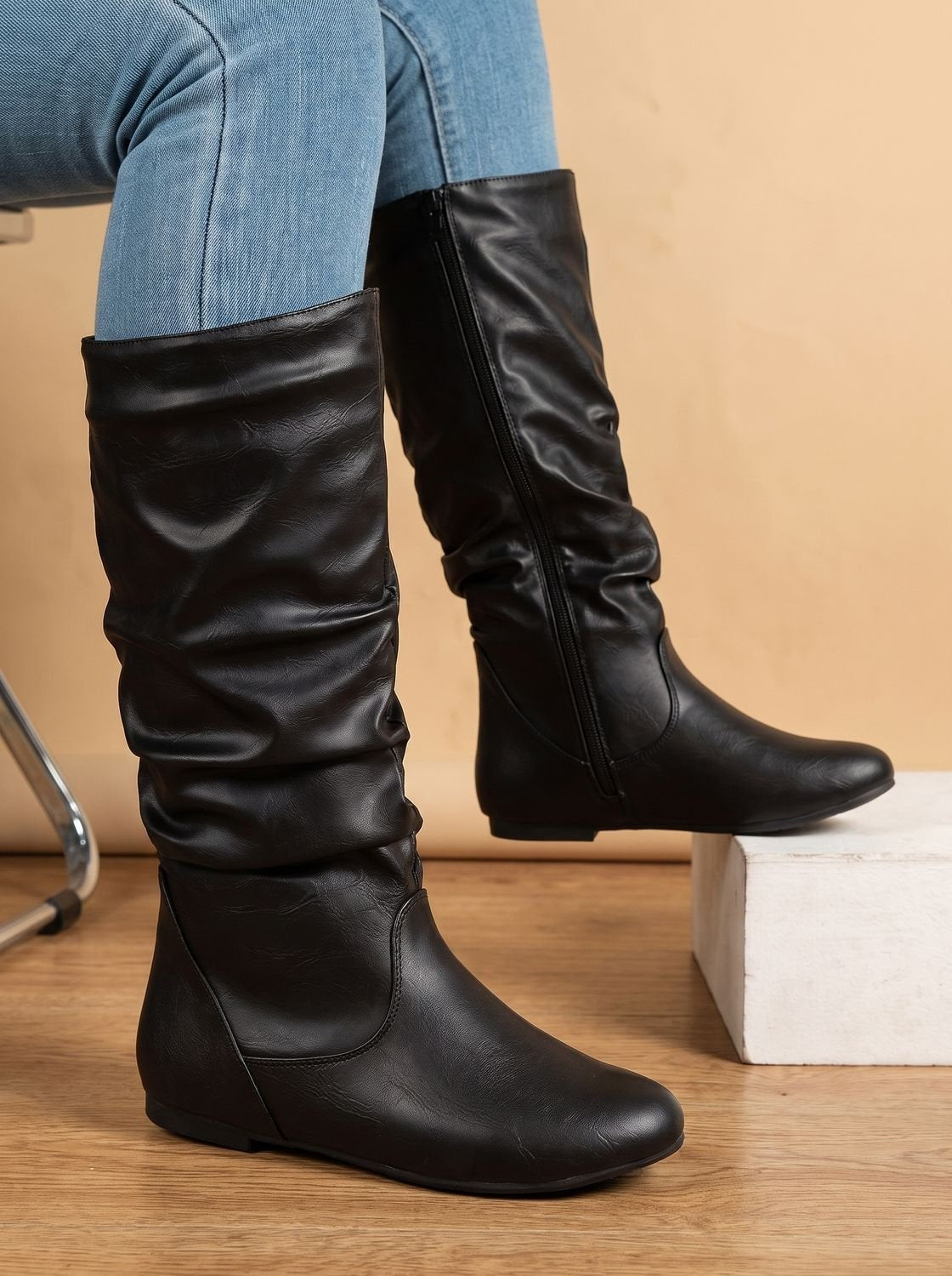 Black knee-high flat boots with gathered shafts and rounded toes, styled with light blue jeans beside a step.