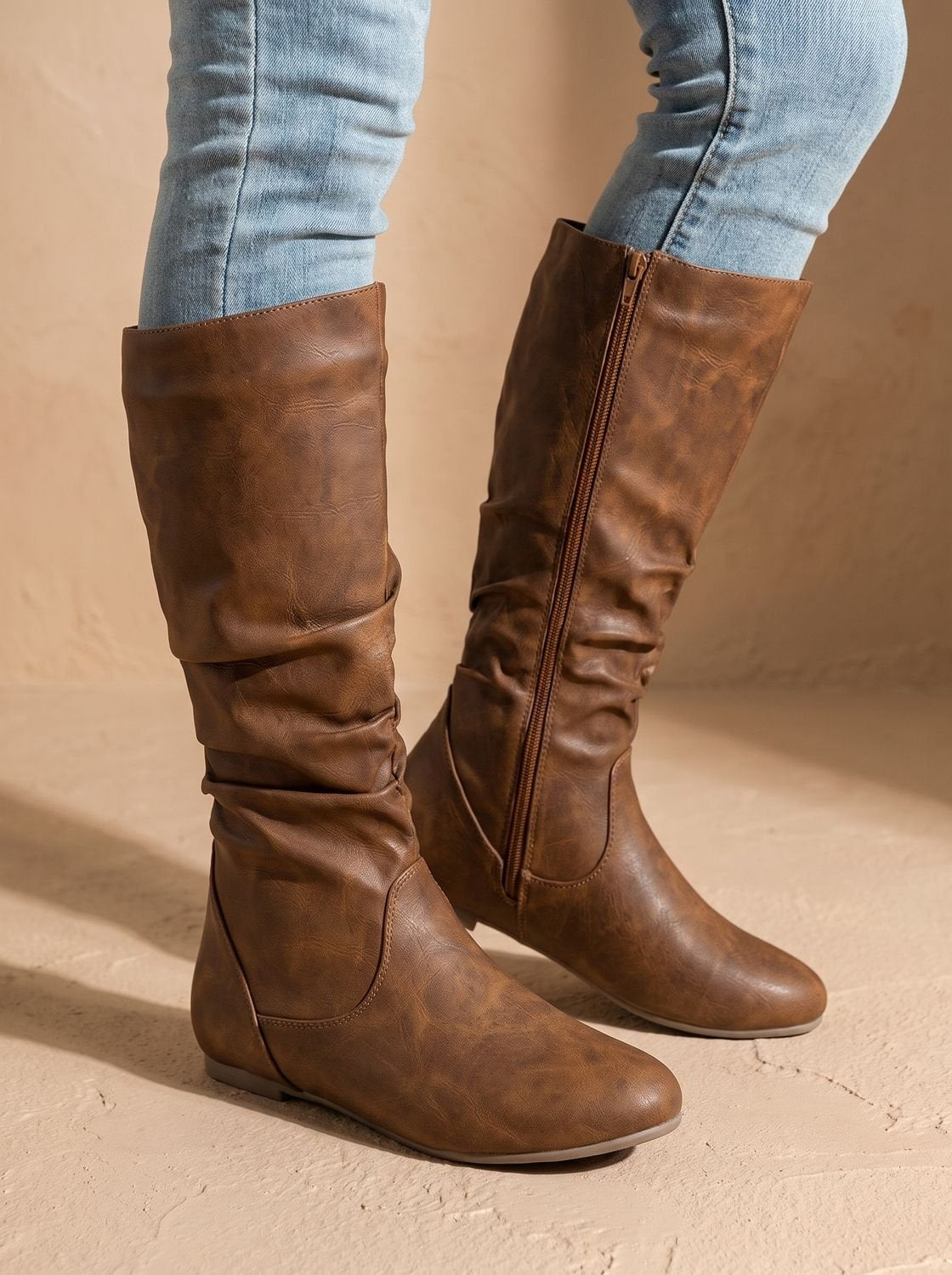 Pair of brown slouchy knee-high boots viewed from the back and side, with full-length zippers and softly gathered shafts.