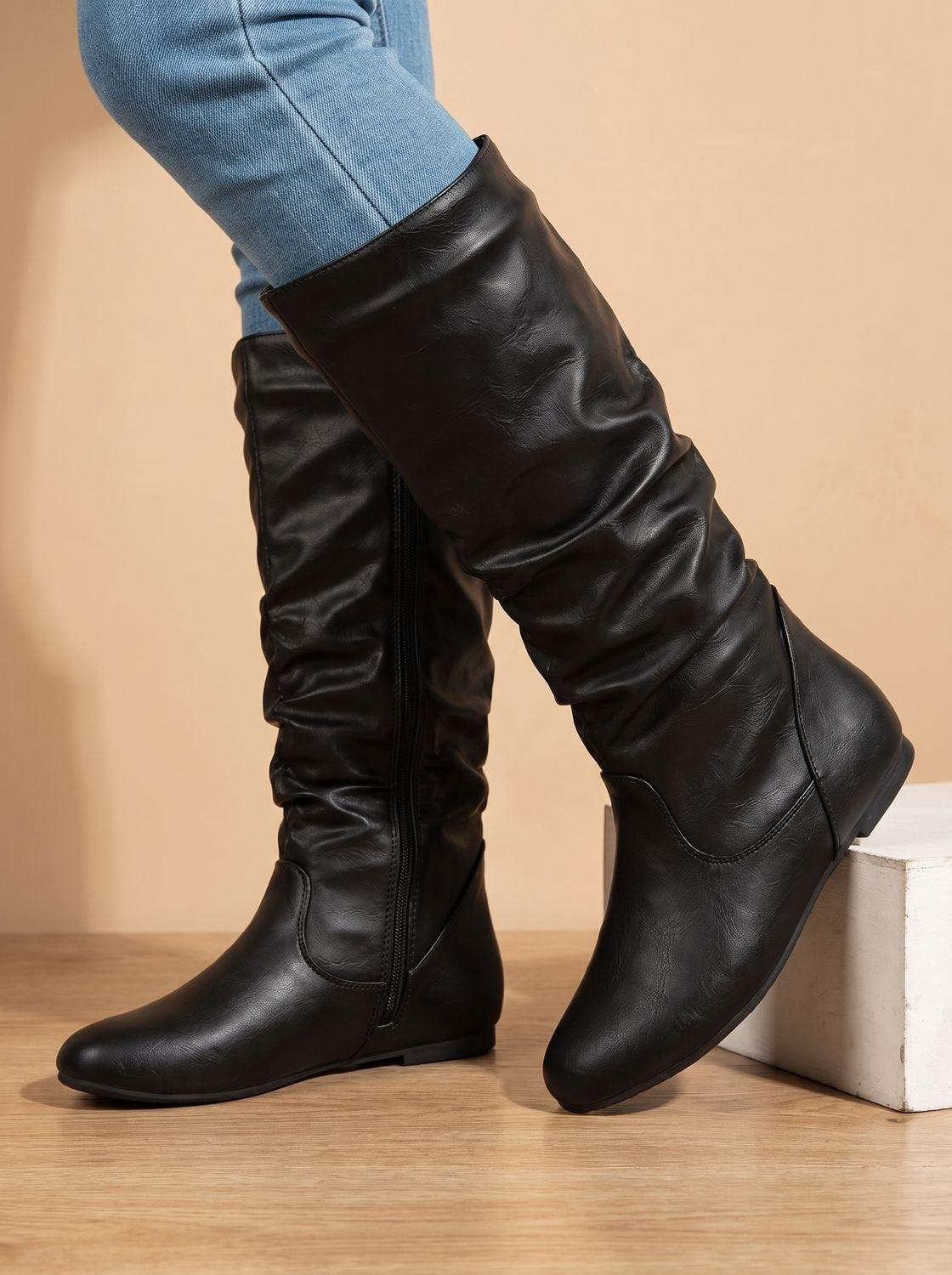 Black slouchy knee-high boot lifted beside a matching boot, emphasizing the gathered shaft, side zipper, and flat sole.
