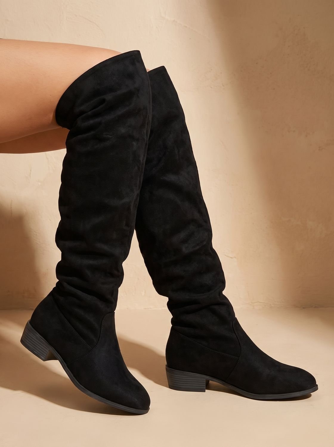 Black slouchy over-the-knee boots worn on crossed legs, highlighting the tall relaxed shafts and pointed toes.