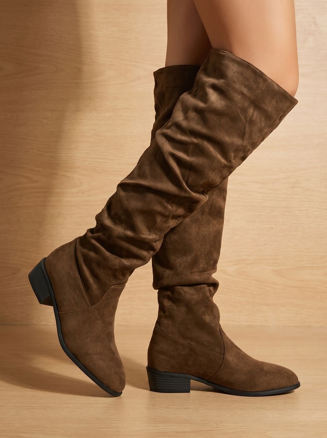 Brown over-the-knee boots on a model in a close side view, emphasizing the layered shaft folds and low heels.