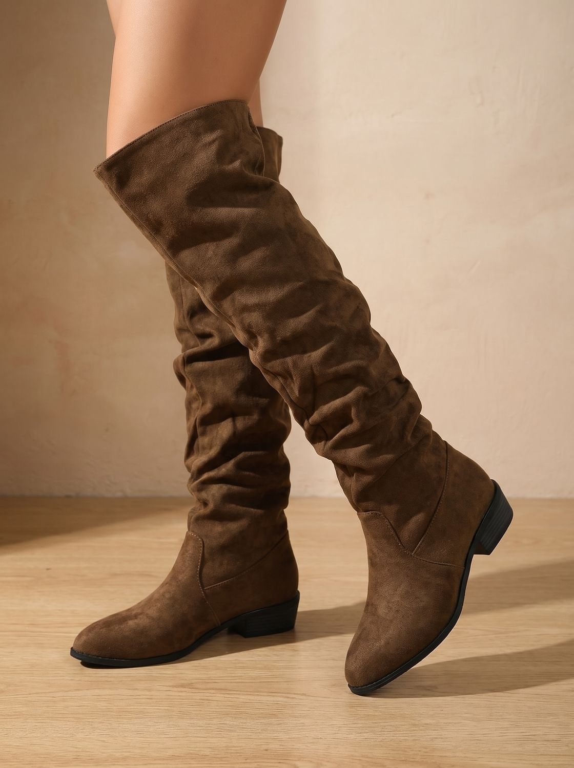 Brown over-the-knee boots with softly ruched shafts, pointed toes, and low heels, shown with one boot lifted.