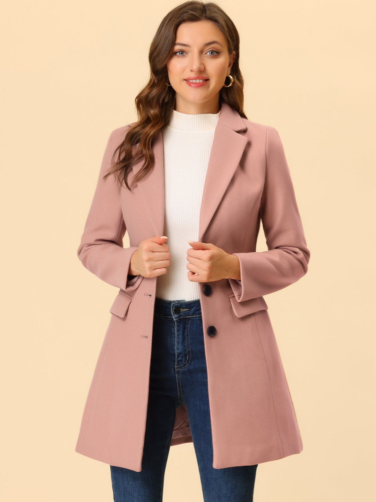 Dusty pink tailored coat worn over a white sweater and jeans, with both hands holding the front.