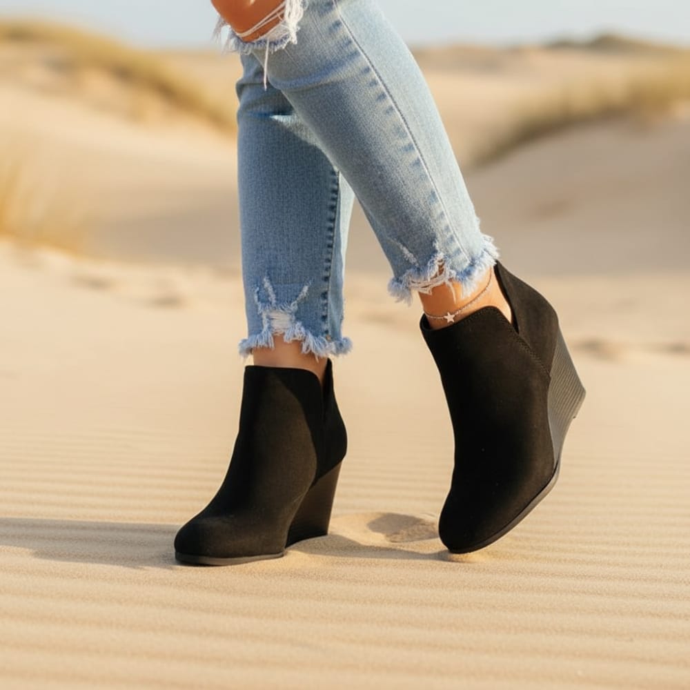 Woman wearing black wedge ankle booties with frayed hem jeans while walking on sandy ground outdoors._0
